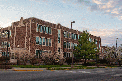 Exterior of a brick, three story building with large windows and trees in front with a blue sky and clouds and the intersection of two roads in front of the building